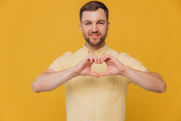 istock Lovely good-looking caucasian bearded guy smiling cheerfully and making heart gesture over chest, expressing his feelings and tenderness while standing against yellow background. I am in love with you 2191659221