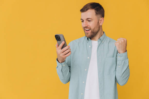 istock Young happy Indian man 30s in basic mint t-shirt hold in hand use mobile cell phone chatting online do winner gesture isolated on yellow background studio People lifestyle concept 2191659974