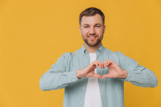 istock Lovely good-looking caucasian bearded guy smiling cheerfully and making heart gesture over chest, expressing his feelings and tenderness while standing against yellow background. I am in love with you 2191660140