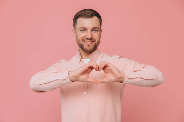 istock Lovely good-looking caucasian bearded guy smiling cheerfully and making heart gesture over chest, expressing his feelings and tenderness while standing against pink background. I am in love with you 2191660384