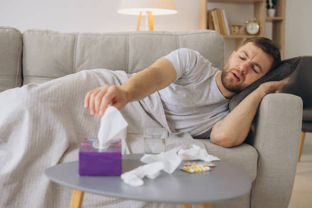 istock Sick caucasian man with a runny nose lying under a warm blanket on the sofa at home and sneezing in paper tissue. Cold, flu, seasonal winter infections concept 2191661871