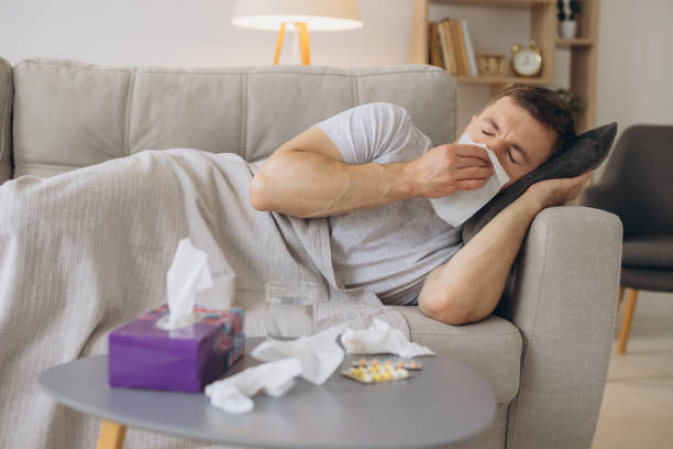 istock Sick caucasian man with a runny nose lying under a warm blanket on the sofa at home and sneezing in paper tissue. Cold, flu, seasonal winter infections concept 2191661874