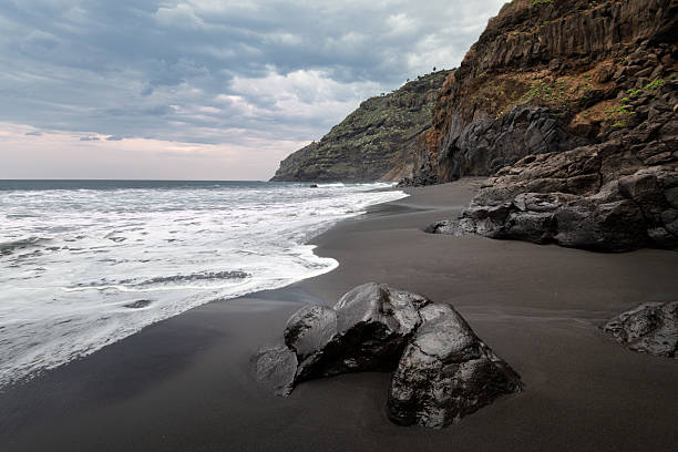 A rocky beach with a black sand and large rock in the foreground. Travel and tourism concept with ocean coastline, Tenerife, Canary Islands.
