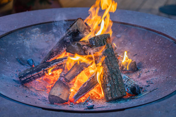 Image depicts a nighttime fire pit, surrounded by burning logs within a circular metal frame The fire shows various stages of combustion and embers No text is present