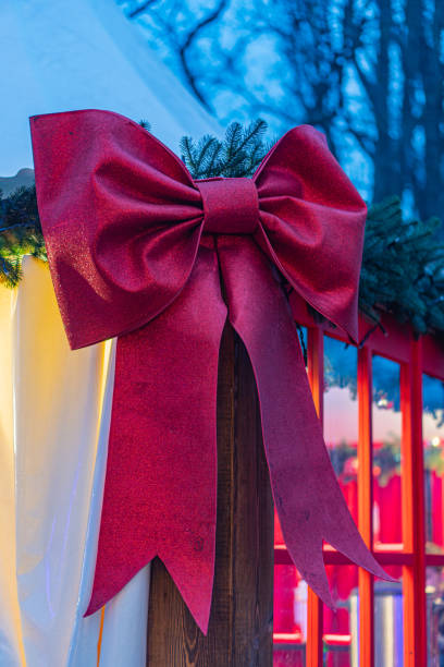 Image showcases a festive red bow with loops and tails, surrounded by pine branches in low light conditions The background is dark, hinting at an outdoor setting The style is realistic and captures
