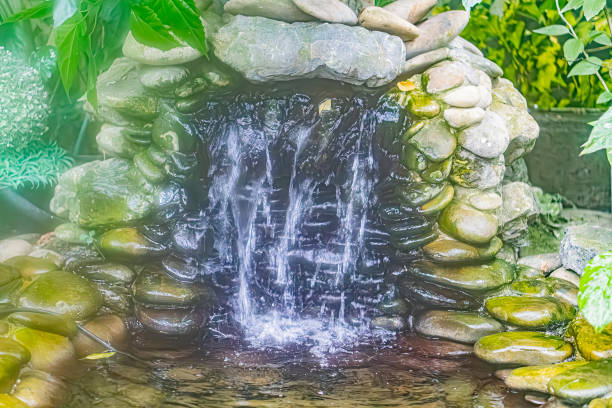 A man-made rock waterfall, partially covered with moss or algae, is set in a lush green garden or park Water cascades from the uneven rocks into a pool below The scene is bathed in soft, diffused l