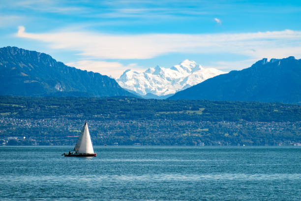 Morges, Switzerland - September 6, 2024: A serene landscape featuring a sailboat gliding across a calm Geneva lake, with snow-capped mountains. The scene is peaceful and idyllic.