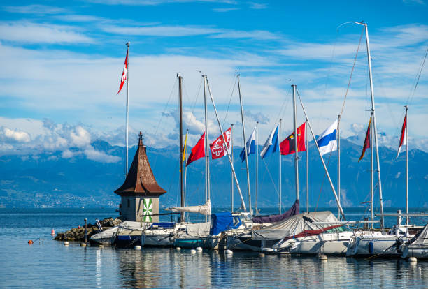 Morges, Switzerland - September 6, 2024: Calm harbor scene with sailboats, moored near charming harbor tower in Morges, Geneva lake. Tranquil waters reflect the scene.