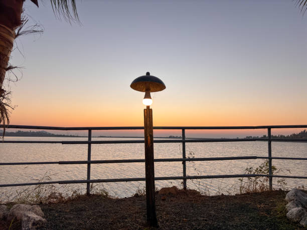 A lighted lamp post is on a pier overlooking a body of water. The sky is a beautiful orange and pink color, creating a serene and peaceful atmosphere