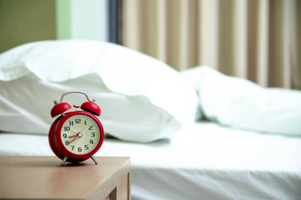 istock Red retro alarm clock on wooden bedside table with white pillows and bedding in softly lit bedroom, evoking calm and cozy morning atmosphere 2191733495