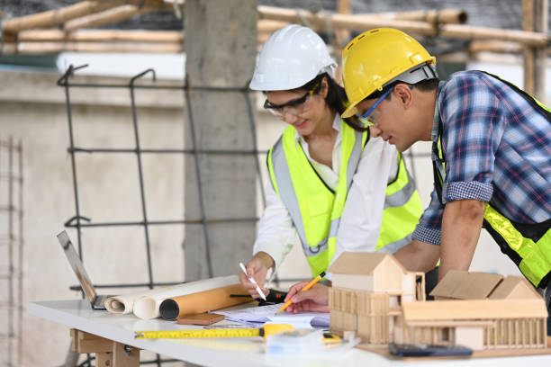 istock Two construction professionals in safety equipment review blueprints and a scale model at a site. They use a laptop and measuring tools, to discuss project details 2191736740