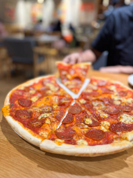 Stock photo of hand holds a delicious slice of thin-crust pepperoni pizza, loaded with savory salami and melted mozzarella cheese. The vibrant Italian restaurant setting adds warmth to this inviting dining moment. Perfectly golden crust, rich toppings, and authentic flavors come together