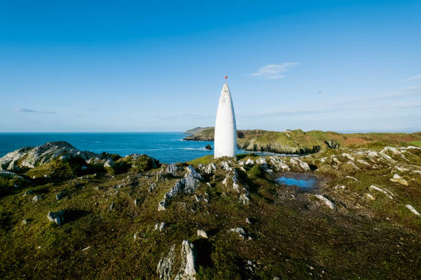 istock Ocean view. A tall, slender, white cone-shaped tower stands prominently on a rocky hill overlooking a tranquil ocean. 2191747074