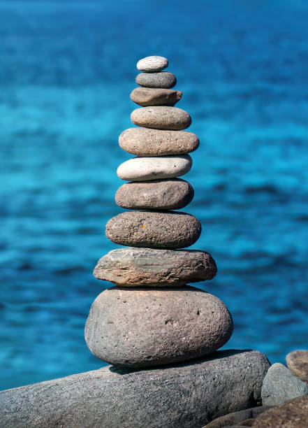 A stone pyramid at Paseo Maritimo cost beach in Adeje, Tenerife with a background of sea and blue sky.A captivating vertical picture of a meticulously balanced stone pyramid at La Caleta coast in Tenerife. Amidst the raw and rugged beauty of the volcanic coastline, the carefully stacked stones stand as a testament to human patience and tranquility. The backdrop of the deep blue ocean adds a serene and meditative quality to the scene.