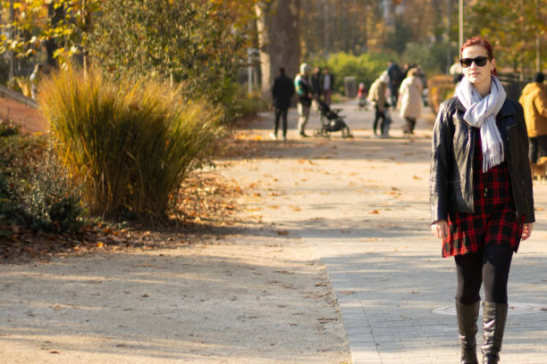 A young woman with red hair and a stylish scarf walks confidently through a sunlit park. The vibrant colors of autumn surround her, creating a lively atmosphere.
