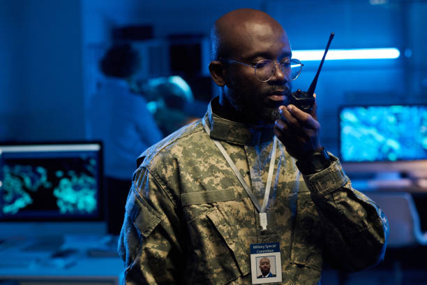 Young African American man in eyeglasses and camouflage uniform speaking in walkie-talkie in front of camera