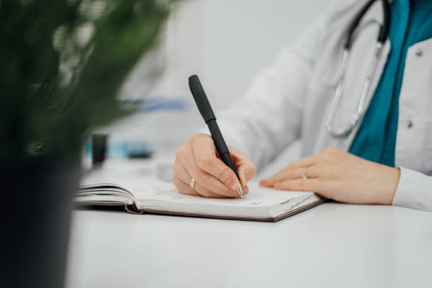 istock Close-up of a doctors hand writing in a notebook with a black pen. A stethoscope is visible on the doctors neck, emphasizing the medical setting. A blurred green plant in the foreground and lab equipment in the background add context and depth to the 2191769228