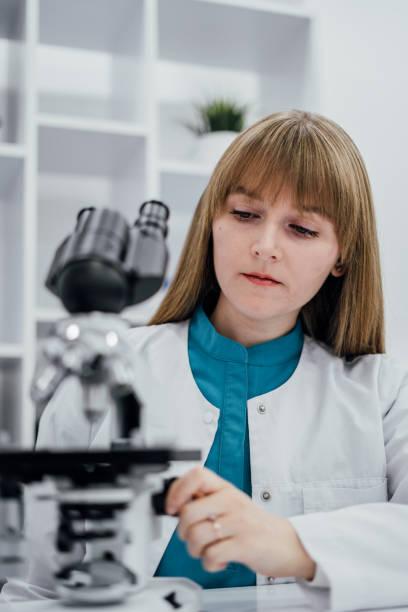 istock A dedicated scientist in a white lab coat and blue uniform is carefully adjusting a microscope to observe samples in detail. The modern laboratory setting with clean shelves and organized tools creates a professional atmosphere. Her focused expression 2191770106