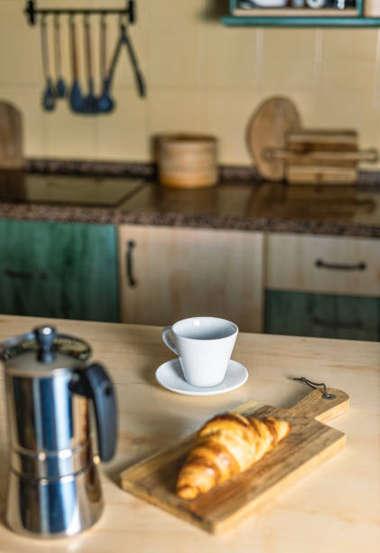 istock Warm light and cozy atmosphere in the kitchen. Coffee pot and hot coffee, empty cup and croissant on a wooden board. 2191778879