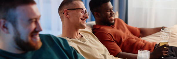 istock Friends Sitting on Sofa Watching TV Smiling Holding Beer Glasses Header Format 2191783895