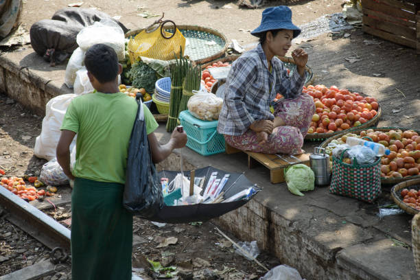 07/12/2019 - Yangon, Myanmar: Two vendors at a Yangon market: a seated woman selling fresh produce such as tomatoes and leafy greens, and a standing man using an umbrella to display goods.
