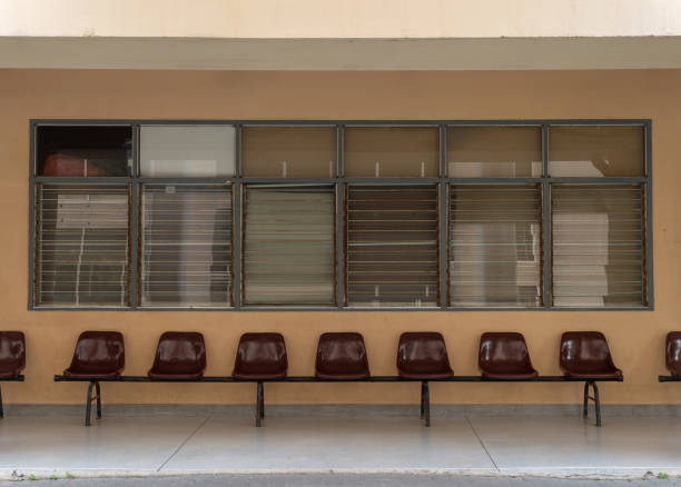 The image shows a wall with several windows and a row of maroon chairs beneath it. The wall is a beige color, and the windows have metal grilles and are partially closed. The chairs are arranged in a straight line, and there are empty spaces between some of them. The overall setting appears simple and unadorned, suggesting a waiting area, possibly in a public building or school.