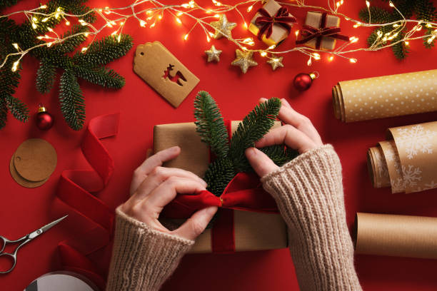 istock Women's hands wrapping a gift. A table with handmade items, rolls of kraft brown paper, red velvet ribbon, labels, scissors. 2191805416