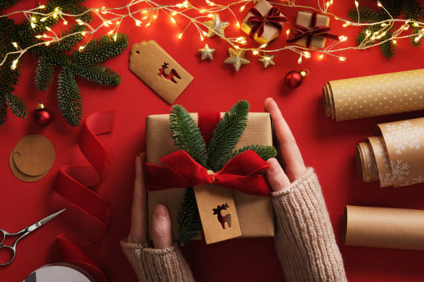 istock Women's hands wrapping a gift. A table with handmade items, rolls of kraft brown paper, red velvet ribbon, labels, scissors. 2191805435