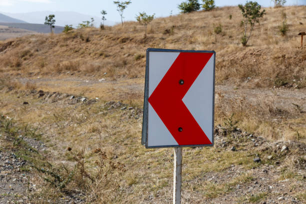 The bright red and white road sign warns drivers of an upcoming sharp left turn in a dry, sparse landscape under a clear sky.