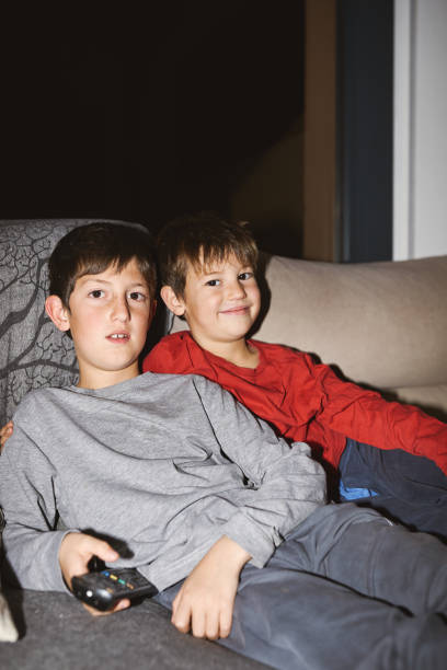 istock Two boys sitting on a couch watching television, one holding a remote control. 2191827380