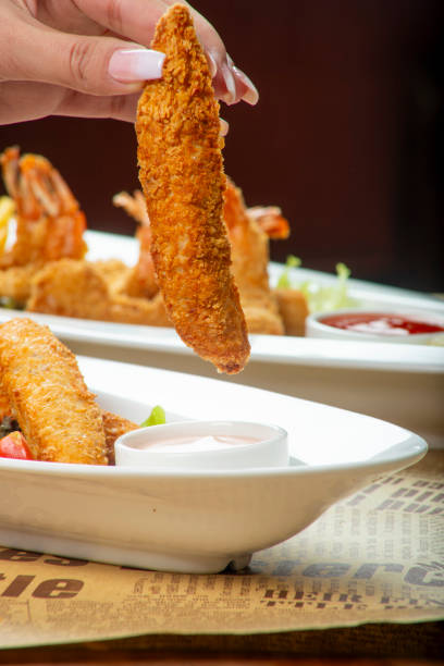 A hand holds a golden brown chicken finger above a plate filled with fried shrimp and fresh vegetables, alongside a small bowl of dipping sauce.
