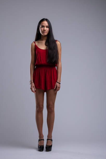 Full-length studio shot of a young woman with long black hair, wearing a red playsuit and black high heels, standing against a plain gray backdrop