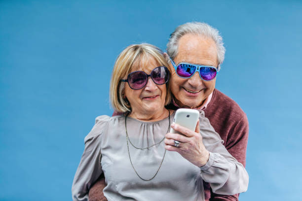 A vibrant senior couple enjoys a playful moment, wearing colorful sunglasses and posing for a selfie against a light blue background. The man, in a burgundy sweater, lovingly embraces the woman, who is dressed in a chic gray blouse accessorized with a necklace. Both radiate happiness and charm as they hold a smartphone, capturing a memory together. Their cheerful demeanor and modern vibe showcase a fun-loving spirit, perfect for celebrating life's moments, anniversaries, or simply enjoying their bond.