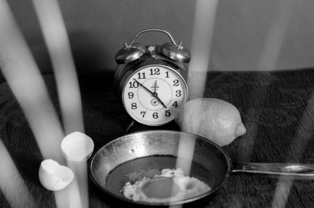 Close-up grayscale still life composition. \n\n\nAn alarm clock sits centrally, with its face visible. The numbers on the clock's face are clearly discernible, and the hands indicate a specific time. \n\n\nNext to the clock, a lemon rests. \n\n\nA small, dark-colored frying pan or skillet is positioned beneath and slightly to the right of the clock, and parts of it extend beyond the edges of the image. \n\n\nTwo pieces of a broken egg are visible, situated near the left edge of the image. \n\n\nThe background is a dark, textured surface, appearing to be a fabric. \n\n\nIntense light streaks or shadows are evident, radiating diagonally across the scene, giving the image a strong sense of directionality. The light creates strong contrasts, highlighting specific parts of the objects.  The overall tone is dramatic and suggests a studio setting.