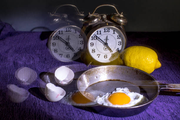 Full indoor shot of a still life arrangement on a purple cloth. In the center, a skillet with a fried egg in it sits on the purple cloth. A lemon rests next to the skillet. Two antique-style alarm clocks are visible, partially overlapping the purple cloth. Several broken egg shells are scattered on the cloth around the skillet and clocks. The time on the clocks is different.
