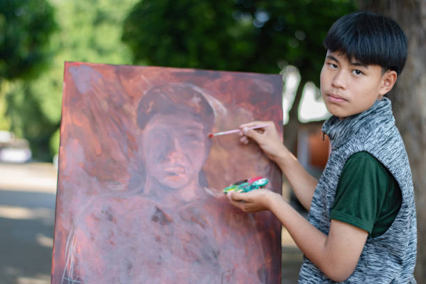 Asian boy holds a paintbrush to paint a human body which is not completed on canvas on a painting frame set in front of him, soft focus, concept for recreational activity and art lesson of children.