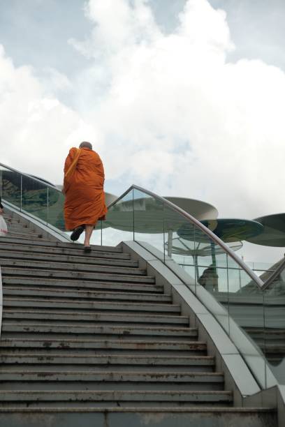Bangkok, Thailand – December 03, 2024: A monk in traditional orange robes ascends modern stairs under a cloudy sky, symbolizing spiritual journey and urban life.