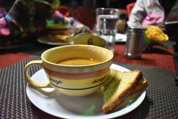 A bowl of warm soup served with a side of toasted bread on a plate. The setting appears to be a restaurant, with other diners visible in the blurred background. The image evokes a feeling of warmth, comfort, and a casual dining experience. The focus is on the soup and bread.