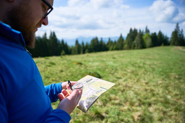 istock Man holds map and compass, standing in green field with trees and mountains in blurred background. 2191868189