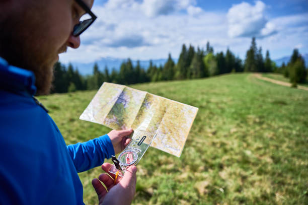 istock Man holds map and compass, standing in green field with trees and mountains in blurred background. 2191868199