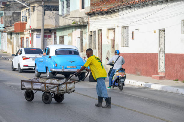 Santa Clara, Cuba - December 26, 2024: A Cuban man pushes a rustic cart in the Central Road. A vintage Chevrolet car is in the background.