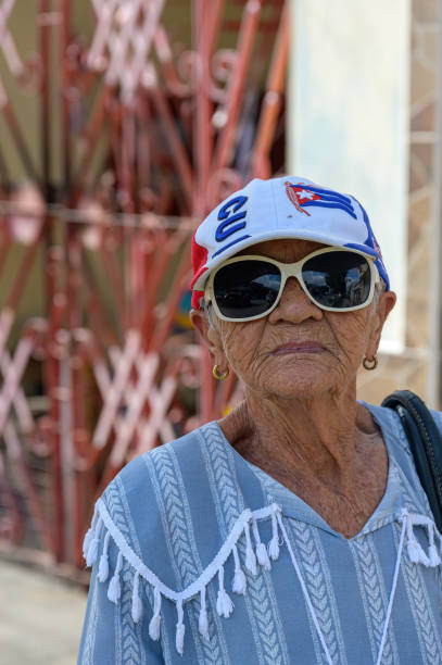 Santa Clara, Cuba - December 24, 2024: Portrait of a Cuban woman wearing a baseball cap with symbols. In the background, security bars in the facade of a house