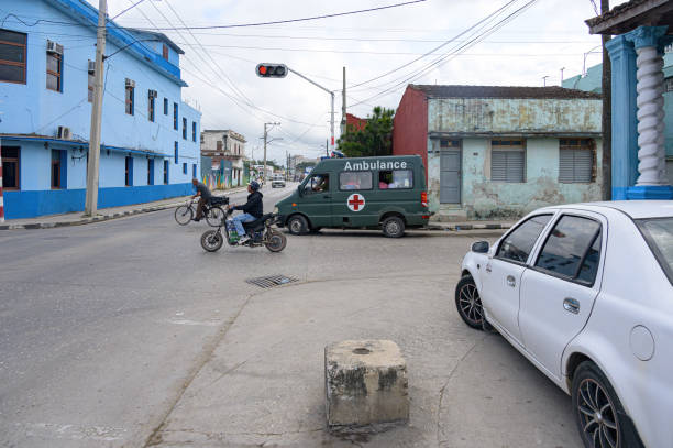 Santa Clara, Cuba - December 28, 2024: An old ambulance full of people drives on an urban road. A man rides a bicycle and another one an electric scooter.