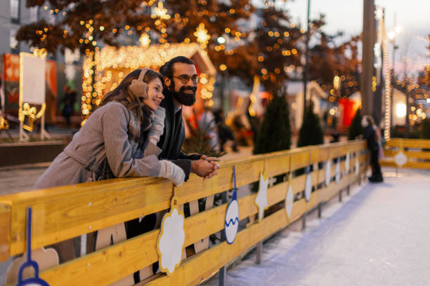 A couple leaning on the rink's railing and watching skaters glide across the ice. They share joyful moment together, surrounded by festive holiday lights and cozy winter atmosphere.