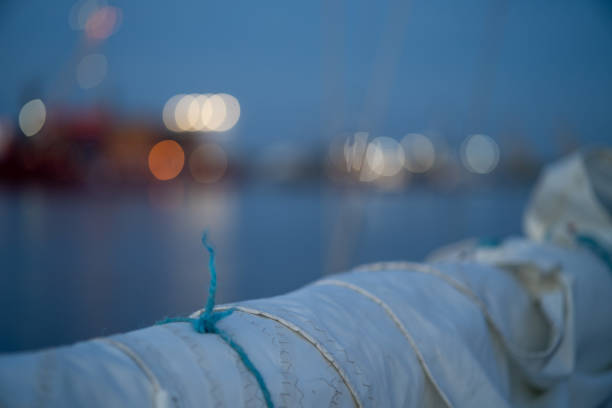 A close-up of a furled sail with a blue rope tied around it, set against a blurred background of harbor lights reflecting on calm evening waters.