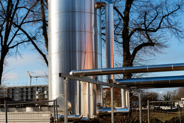 A large metallic storage tank stands beside a network of pipes on an industrial site. The structure reflects sunlight, while nearby trees and a construction site complete the backdrop.