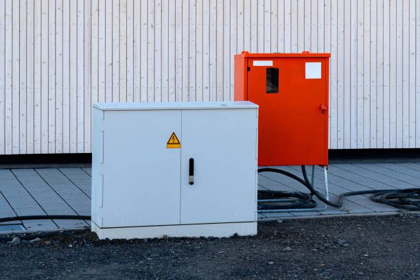 A white utility box and an orange control box are positioned on gravel near a wooden building. They serve essential functions in managing electrical connections and equipment.