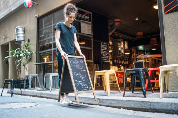 istock Determined Young Waitress Arranges Sidewalk Chalkboard Menu At Urban Cafe 2191960613