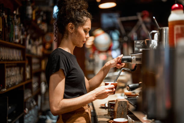 Aboriginal waitress in black attire operates professional espresso machine, demonstrating skilled coffee preparation techniques in a warmly lit cafe environment with traditional bar setup.