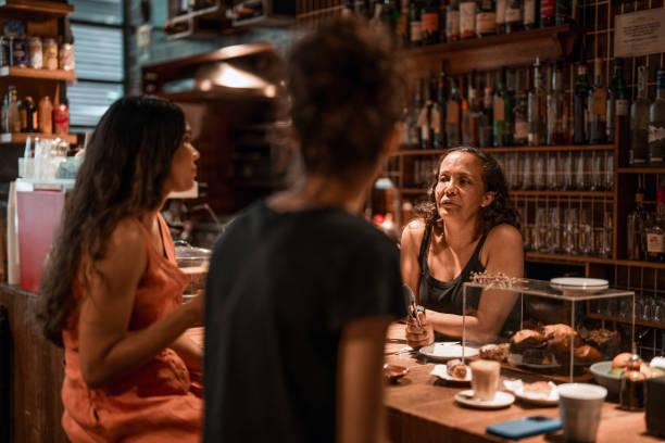 Indigenous female customers engage with professional server at wooden bar counter, surrounded by bottles in warm cafe lighting.